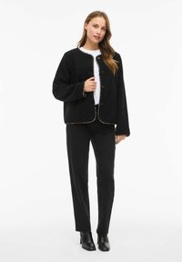 Giacca in pile nera con finitura testurizzata, scollatura tonda, bottoni frontali e rifinitura con motivo leopardato. Abbinata a pantaloni neri e stivali con tacco.