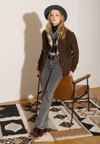 Veste en daim marron à franges, col roulé noir, chemise à carreaux, jean évasé gris et bottines bordeaux, accessoirisés d'un chapeau et d'une écharpe gris.