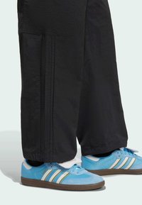 Niebieskie buty sportowe z trzema białymi i złotymi paskami, w połączeniu z czarnymi, luźnymi spodniami z kieszenią boczną i fakturą.