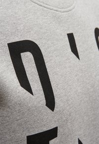 Gros plan sur un tissu gris avec de grandes lettres noires partielles imprimées à la surface, montrant la texture et les coutures autour de l'encolure.