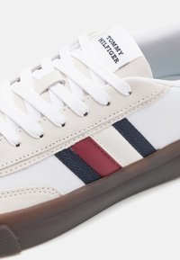 Baskets blanches avec détails en daim beige, bandes bleu marine et rouge sur le côté, lacets blancs et étiquette "Tommy Hilfiger" sur la languette.