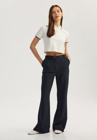Witte cropped polo shirt met zwarte accenten, gecombineerd met donkerblauwe verticaal gestreepte wijde broek. Model staat met handen in de zakken.