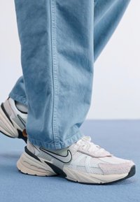 Sneaker Nike bianchi e grigi con accenti in mesh e suede, dotate di una suola curva e un logo Swoosh nero, abbinate a pantaloni tapering blu chiaro.