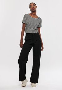 Zwarte wijde broek gecombineerd met een zwart-wit gestreept T-shirt met korte mouwen. Schoenen zijn lichtbeige sneakers. Glad materiaal en casual pasvorm.