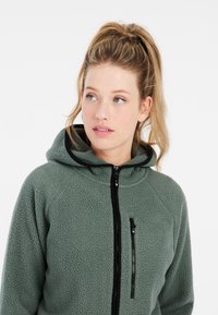 Groene fleecetrui met een gestructureerde afwerking, voorzien van een zwarte rits, een verstelbare capuchon en een ritszak op de borst.