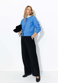 Cardigan tricoté bleu clair à col rond, pantalon noir à jambes larges et chaussures noires à motifs. Modèle posant devant un fond blanc.