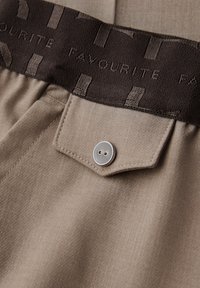 Gros plan sur un pantalon en tissu beige avec une poche à rabat boutonnée et une ceinture noire portant l'inscription "FAVOURITE" en lettres majuscules.