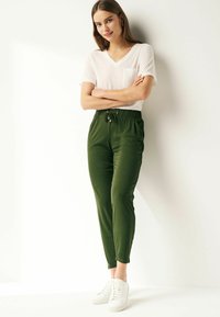 Grønne joggerbukser med elastisk talje og lommer, parret med en hvid V-hals T-shirt. Model iført hvide sneakers, stående mod en lys baggrund.