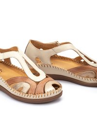 Sandalias de cuero marrón y crema con punta abierta, acentuadas por costuras decorativas y detalles recortados. Plantilla texturizada y suela marrón.