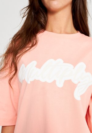 Femme aux longs cheveux bruns portant un T-shirt rose clair avec du texte en cursive blanche "Multiplier" sur la poitrine.