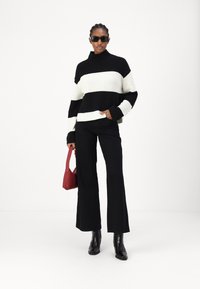 Maglione a righe bianche e nere in maglia con collo alto, abbinato a pantaloni neri a zampa d'elefante e stivali neri. In mano tiene una piccola borsa rossa.