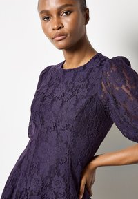 Fioletowa sukienka z bufkami wykonana z koronki, ozdobiona kwiatowymi wzorami i jednolitą linią dekoltu. Zaprojektowana w formie strukturalnej z teksturowanym detalem.