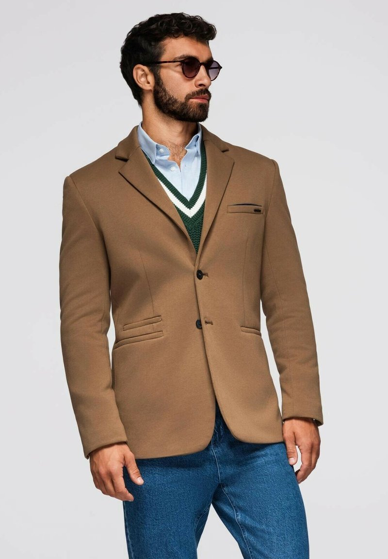Blazer marrón con un corte ajustado, cierre de un solo botón y dos bolsillos frontales, llevado sobre un chaleco a rayas verdes y blancas y unos vaqueros azules.