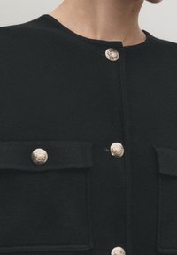 Svart plagg med silverfärgade dekorativa knappar, horisontella fickor med lock och en slät tygtextur. Rund halsringning, figursydd design.