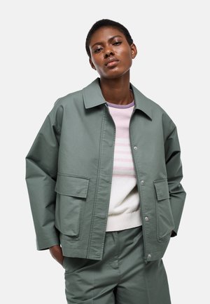 Femme portant une veste et un pantalon vert sauge avec un pull rayé blanc et rose, posant devant un fond clair uni.