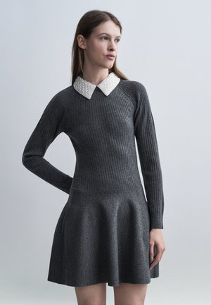 Femeie purtând o rochie gri închis cu mâneci lungi, cu textură nervurată, cu fustă evazată și un guler alb decorat cu perle, stând pe un fundal gri simplu.