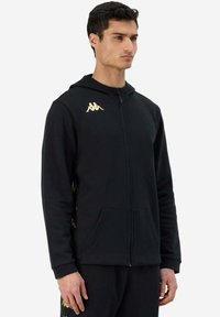 Felpa nera con zip realizzata in tessuto morbido, con un logo dorato sulla parte sinistra del petto e dettagli dorati lungo i lati. Include una tasca frontale.