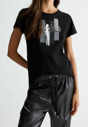 Kobieta nosząca czarny T-shirt z graficznym abstrakcyjnym wzorem oraz czarne spodnie z ekoskóry ze ściągaczem w pasie i bocznymi kieszeniami.