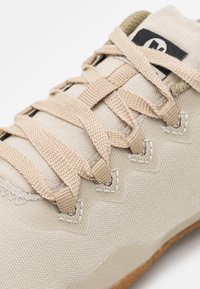 Beige tygsko med strukturerad yta, förstärkta sömmar, platta skosnören och gummisula. Har öljetter för justering av snörningen.