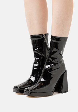 Bottines en cuir verni noir avec fermeture éclair à l'avant, bout carré et talon large, présentant une finition brillante et un design épuré.