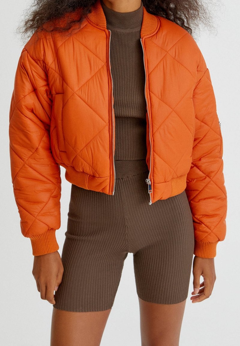 Blouson aviateur matelassé orange avec col et poignets côtelés, fermeture éclair. Associé à un short marron côtelé ajusté.