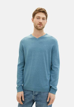Pull en V en tricot bleu doux, avec des manches longues et des poignets et ourlet côtelés, porté avec un jean bleu clair. Logo subtil sur la poitrine.