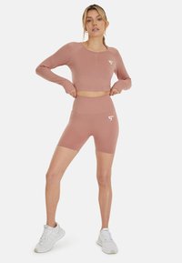Roze lange mouwen crop top en bijpassende high-waisted shorts van rekbaar materiaal, met witte logo-accenten. Draagt met witte sneakers.