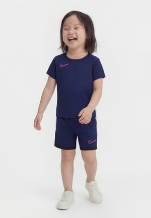 Smilende barn med mørkt hår iført mørkeblå Nike-sportsbeklædning og hvide sneakers, stående på en ensfarvet hvid baggrund.