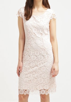 Vrouw die een knie-lange, kortemouwige, lichtroze kanten jurk draagt met cirkelvormige bloemmotieven op een witte achtergrond.