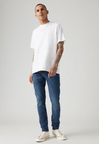 Weißes Baumwoll-T-Shirt mit lockerer Passform, kombiniert mit blauen Jeans und weißen Sneakern mit roten Akzenten und strukturierten Schnürsenkeln.