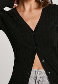 Černý žebrovaný cardigan s V-výstřihem a modrými knoflíky. Má přiléhavý střih a dlouhé rukávy, v kombinaci se šedými džíny s vysokým pasem.