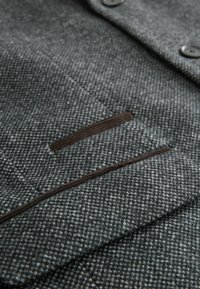 Grå stof med et struktureret houndstooth-mønster, der har to mørke knapper og en kantet lomme detalje. Blød, vævet tekstur.