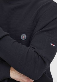 Maglione crewneck blu in tessuto testurizzato, con una patch rotonda del logo sul petto e un'accente tricolore sulla manica.