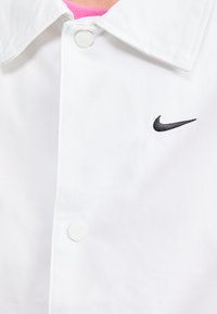 Bijela košulja s preklopnim ovratnikom, s crnim Nike swoosh logotipom izvezenim s lijeve strane i bijelim gumbom na pritiskanje.