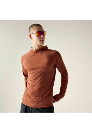Mann trägt ein oranges langärmliges Sportshirt und reflektierende Sportsonnenbrille, steht mit den Händen hinter dem Rücken vor einfarbigem Hintergrund.