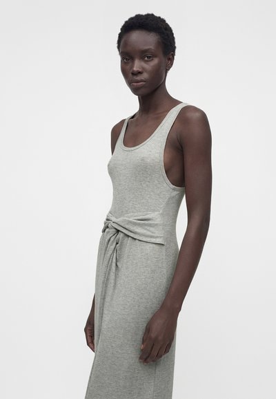 Λεπτή μαύρη γυναίκα με κοντά μαλλιά, που φοράει ένα γκρι αμάνικο φόρεμα δεμένο με κόμπο στη μέση, στέκεται μπροστά σε ένα απλό ανοιχτό φόντο.