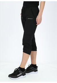 Pantaloni tuta neri corti con una vita elasticizzata, tasche laterali e dettagli con logo. Indossati con sneakers nere con suola bianca.