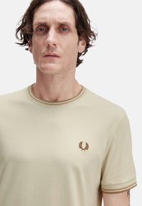 Camiseta de color beige claro con mangas cortas, que presenta un cuello acanalado y un logo marrón bordado en el pecho. Textura de algodón suave.