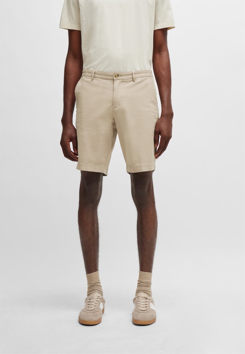 Shorts de algodón beige con un corte ajustado, que presentan bolsillos laterales, cierre frontal con botón y dobladillos enrollados. Llevados con zapatillas de color claro.