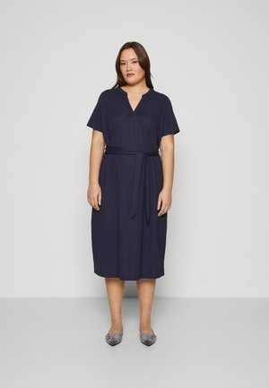 Navy blue knee-length dress in lightweight fabric, featuring short sleeves, a v-neck, and a tied belt at the waist. Silver flat shoes.
