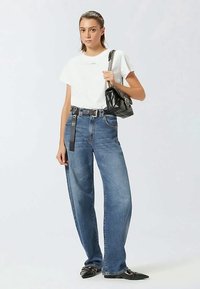 Young woman wearing white t-shirt, high-waisted blue jeans, black belt, black pointed shoes, holding black quilted handbag on shoulder.