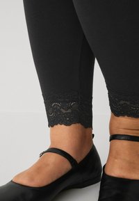 Zwarte leggings met kant aan de onderkant, gecombineerd met zwarte platte schoenen met een bandje over de enkel.