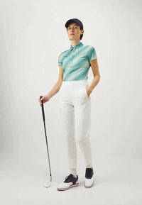 Golfový outfit pozostávajúci z tyrkysového polo trička so vzorom, bielych elegantných nohavíc a námorníckej čiapky. Doplnky zahŕňajú golfovú palicu a topánky s bielymi akcentmi.