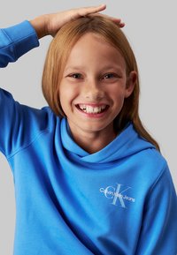 Sudadera de algodón azul con diseño de cuello de cowl. En la parte delantera tiene un logo blanco de Calvin Klein Jeans. Textura suave, corte relajado, mangas largas.