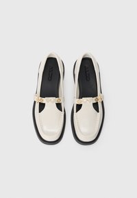 Mocassins en cuir verni blanc avec une semelle noire, mettant en avant une sangle décorative avec des accents dorés et des détails en forme de cœur.