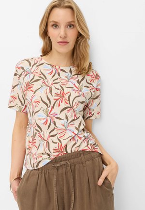 Kvinde med skulderlangt blondt hår iført en beige kortærmet skjorte med blomsterprint og brune bukser med snøre i taljen, hænderne i lommerne.