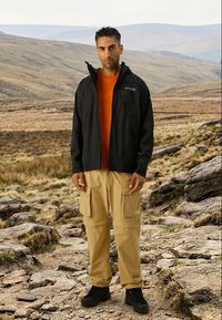 Férfi fekete dzsekiben, narancssárga ingben, bézs cargo nadrágban és fekete bakancsban, aki egy sziklás ösvényen áll egy dombos, szabadtéri környezetben.