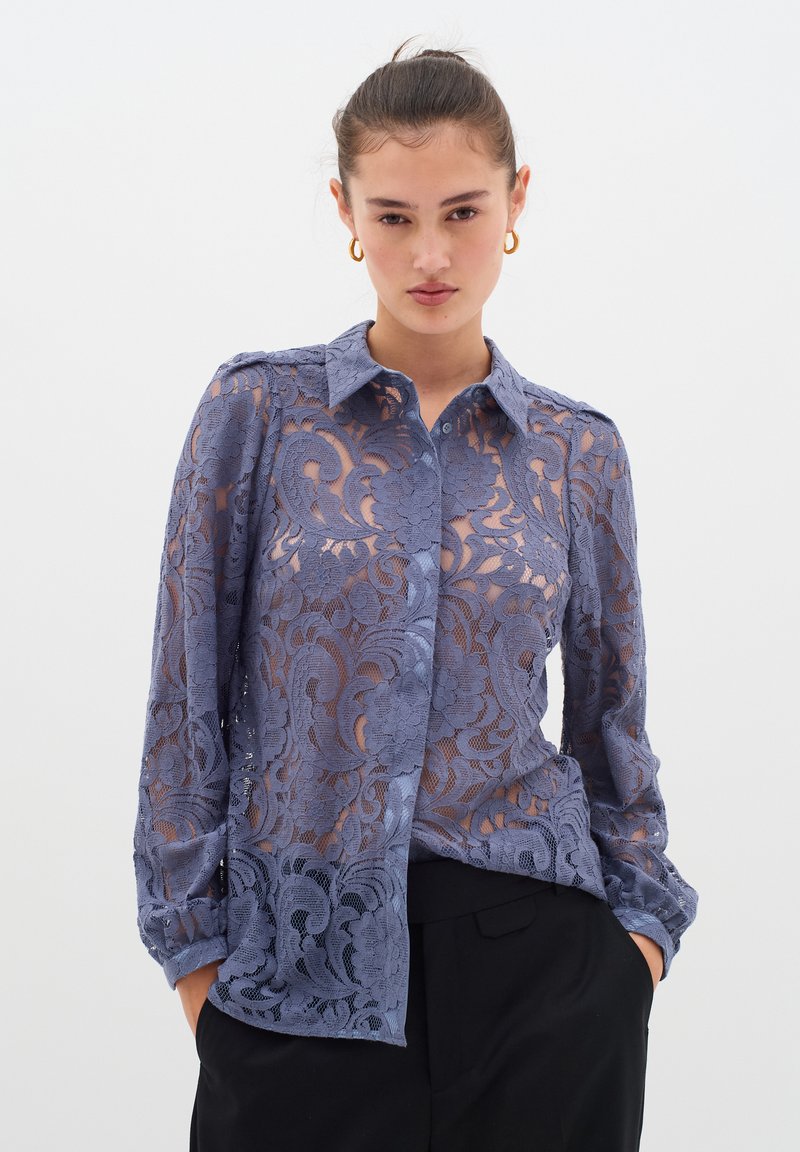 Cămașă din dantelă de un albastru deschis, cu mâneci lungi, guler și modele florale. Textura transparentă dezvăluie stratul de dedesubt. Pantaloni negri purtați dedesubt.