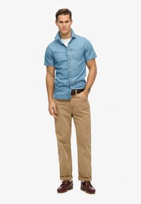 Jasnoniebieska koszula z chambray, z krótkim rękawem, zapinana na guziki, w połączeniu z khaki spodniami i brązowymi butami typu lace-up. Posiada kieszonkę na piersi.