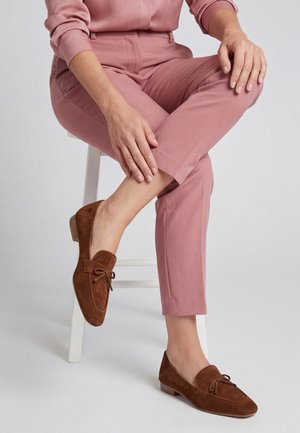 Osoba siedzi na białym stołku, ubrana w różowe spodnie i brązowe zamszowe mokasyny z małymi kokardkami, dłonie spoczywają na skrzyżowanej nodze.